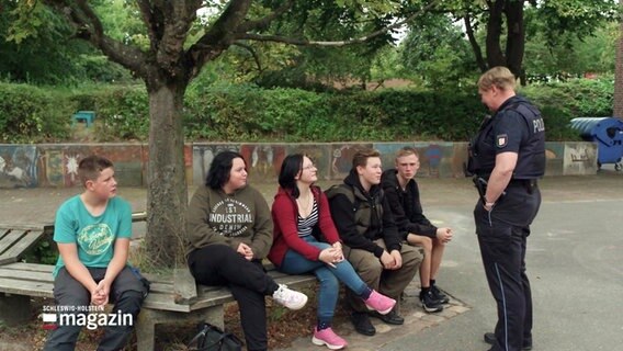 Eine Polizistin spricht auf einem Schulhof mit einer Gruppe von Schülerinnen und Schülern. © Screenshot 