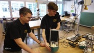Zwei junge Männer stehen in einem Klassenraum und präsentieren eine eigene Innovation. © Screenshot 