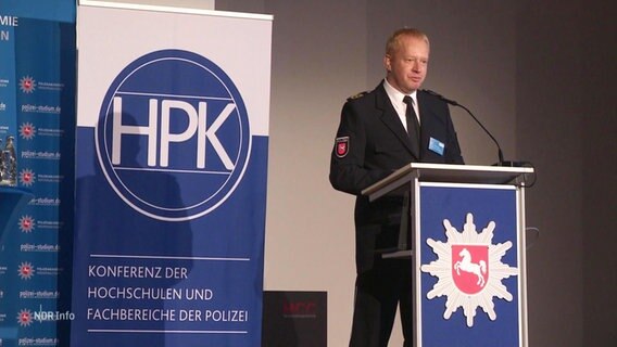 Ein Sprecher steht bei einer Konferenz der Hochschulen und Fachbereiche der Polizei auf einer Bühne und hält eine Rede. © Screenshot 