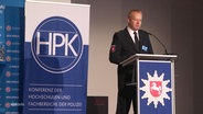 Ein Sprecher steht bei einer Konferenz der Hochschulen und Fachbereiche der Polizei auf einer Bühne und hält eine Rede. © Screenshot 