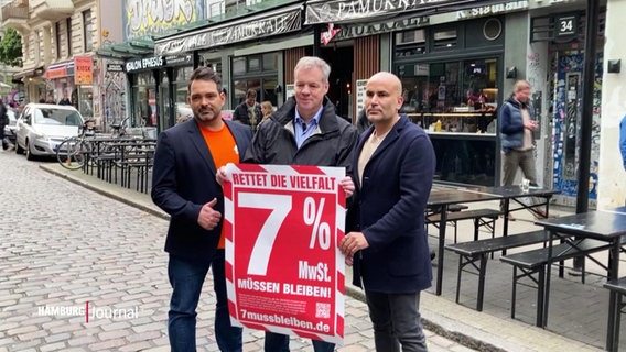 Drei Gastronomen stehen im Schanzenviertel mit einem Plakat mit der Aufschrift "7% Mehrwertsteuer müssen bleiben". © Screenshot 