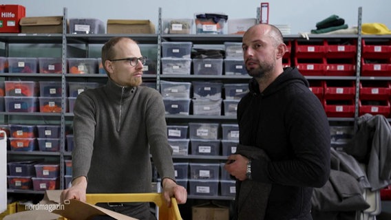 Zwei Männer stehen vor einem Regal mit Plastikboxen: Marco Scheel, Gründer und Geschäftsführer von Nordwolle Rügen und sein Mitarbeiter Nicolai Zuicov. © Screenshot 