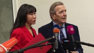 Altkanzler Gerhard Schröder und seine Frau So-yeon Schröder-Kim vor Mikrofonen. © Screenshot 