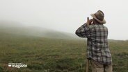 Ein Mann mit kariertem Hemd, Hut und Wanderstock von hinten zu sehen, der die Hände zum Rufen vor den Mund hält. Er steht auf einer steil ansteigenden Almwiese. © Screenshot 