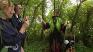 Eine als Hexe verkleidete Frau mit einer grünen Langhaar-Perücke streckt theatralisch die Arme aus in einem Wald. © Screenshot 