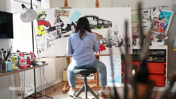 Eine Frau sitzt auf einem Hocker vor einer bunt bemalten, größeren Leinwand in einem Atelier. © Screenshot 
