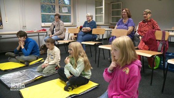 Mehrere Kinder sitzen auf gelben Matten vor ihren Großeltern bei einem Erste-Hilfe-Kurs. © Screenshot 