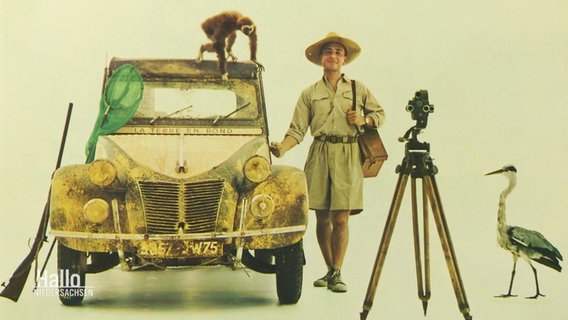 Ein Mann im "Dschungel-Safari"-Outfit steht im Fotostudio neben einem Auto des Modells "Ente", neben ihm ein Kamerastativ, ein Kranich und ein kleinerer Affe. © Screenshot 