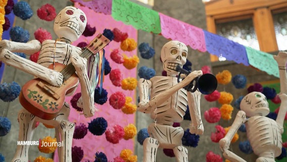 Skelett-Figuren mit Instrumenten stehen vor einer bunten Fassade. © Screenshot 