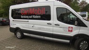 Ein weißer Kleinbus mit der Aufschrift "CariMobil" steht auf einem Parkplatz. © Screenshot 
