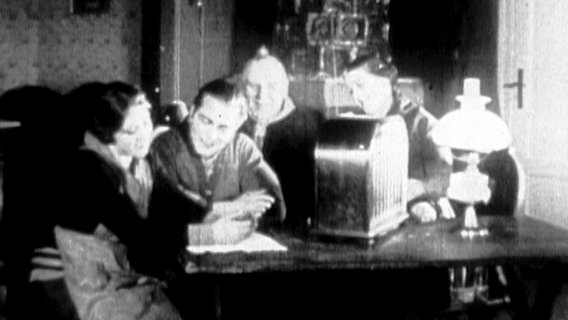Ein altes Schwarz-Weiß-Foto zeigt eine Gruppe von Menschen, die Radio hören. © Screenshot 