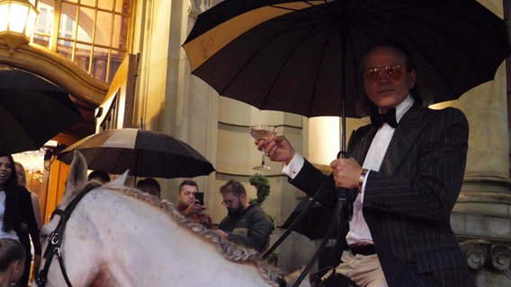 Eine Person mit Regenschirm und Glas in den Händen auf einem Pferd. © Screenshot 