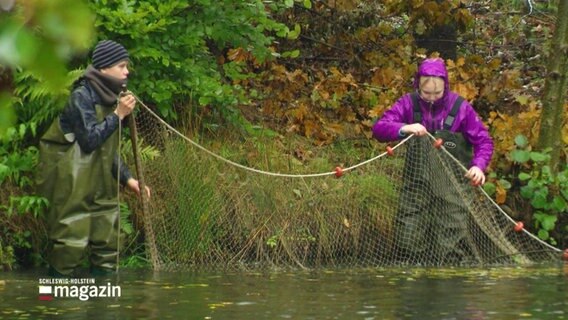 Zwei Personen mit Regenkleidung stehen mit einem Fischernetz im Wasser. © Screenshot 