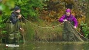 Zwei Personen mit Regenkleidung stehen mit einem Fischernetz im Wasser. © Screenshot 