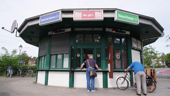 Ein freistehender, vieleckig gebauter Kiosk. Ein Mann steht am Ladenfenster und bestellt etwas, eine Person stellt währenddessen gerade ein Fahrrad ab. © Screenshot 