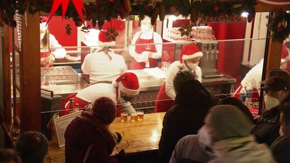 Ein weihnachtlich geschmückter Stand auf einem Weihnachtsmarkt; die Mitarbeitenden tragen Nikolausmützen und rote Schürzen über weißen Pullovern. © Screenshot 