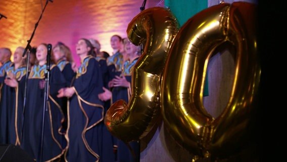 Im Bildvordergrund rechts goldene Folienballons, die die Zahl 30 bilden, unscharf im Hintergrund ein Gospelchor. © Screenshot 