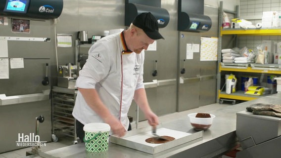 Ein Mann steht in einer Großküche und verteilt flüssige Schokolade in eine Form. © Screenshot 