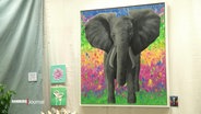 An einer Wand hängt ein größeres Gemälde von einem Elefanten vor einem bunten Hintergrund. © Screenshot 