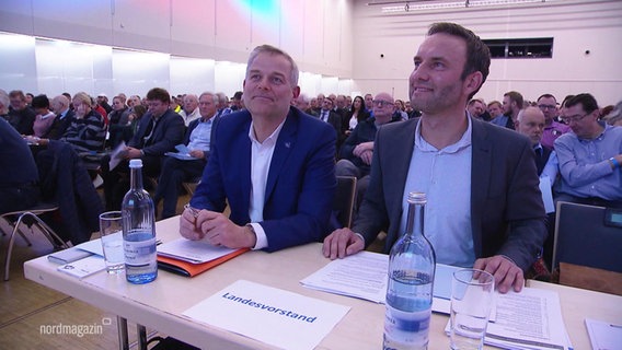 Leif-Erik Holm und Enrico Schult sitzen mit zufriedenem Lächeln in der ersten Reihe auf einem Landesparteitag. © Screenshot 