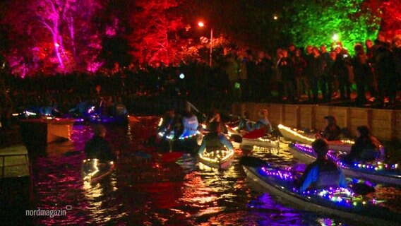 Bei einem Lichterpaddeln fahren mit bunten Lichterketten behangene Kanus durch kleine Kanäle an Zuschauenden vorbei. © Screenshot 
