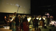 Ein Orchester ist vor einer Leinwand positioniert. © Screenshot 