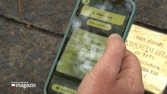 Eine Hand mit einem Smartphone, auf dem Display sieht man das Bild eines Stolpersteins und weiterführende Informationen. Im Bildhintergrund ein Stolperstein im Gehweg. © Screenshot 