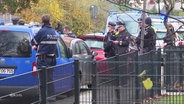 Szene auf einer Straße: Polizisten in Uniform haben eine Straße abgesperrt, mehrere Beamte im Bild, einer von ihnen telefoniert. © Screenshot 