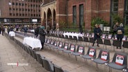 Vor dem Haupteingang zur Petrikirche sind 200 Stühle in Reihen aufgestellt, an jedem Stuhl klebt ein Foto und ein Name einer Geisel der Hamas in Israel. Bürgerinnen und Bürger gehen zwischen den Stuhlreihen hindurch. © Screenshot 
