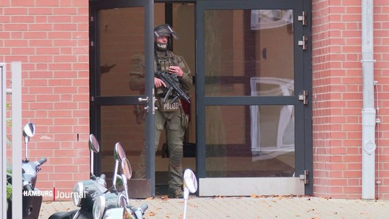 Ein Polizist in olivgrüner Kampfmontur steht in der Eingangstür einer Schule, um den Hals hängt ein Maschinengewehr. © Screenshot 