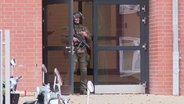 Ein Polizist in olivgrüner Kampfmontur steht in der Eingangstür einer Schule, um den Hals hängt ein Maschinengewehr. © Screenshot 