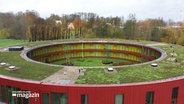 Aus der Vogelperspektive: Ansicht des Internats in Güby. Ein rund erbautes, zweigeschössiges Gebäude mit Gras auf dem Dach, einem begrünten Innehof und einer roten Fassade. © Screenshot 