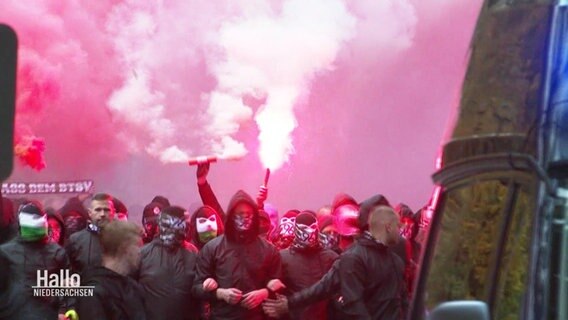 Eine Gruppe von maskierten zieht durch eine Straße, zündet Pyrotechnik und trägt Banner bei sich. © Screenshot 