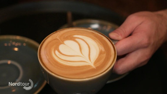 In einer Cappuccino-Tasse ist eine Blume mit Milchschaum gemalt. © Screenshot 