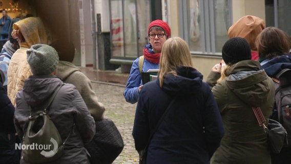 Eine kleinere Gruppe von Zuhörenden steht um eine erzählende Frau in einer Fußgängerzone bei einer Stadtführung. © Screenshot 