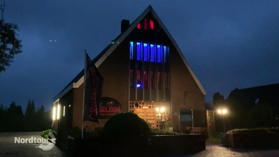 Blick bei Nacht auf ein modernes, ehemaliges Kirchengebäude das nun als Gruselmuseum bunt beleuchtete Fenster aufweist. © Screenshot 
