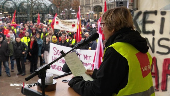 Eine Frau in gelber Warnweste mit dem ver.di-Logo darauf spricht bei einer Demonstration auf einer Bühne in ein Mikrophon. © Screenshot 
