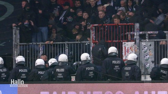 Eine Gruppe von Polizisten stehen in Schutzausrüstung inklusive Helm vor einem Fanblock im Fußballstadion. © Screenshot 