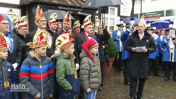Moritz Enneking (rechts im Bild), Präsident Dammer Carnevalgesellschaft, hält eine Rede- Links von ihm stehen zahlreiche Menschen mit Narrenkappen. © Screenshot 