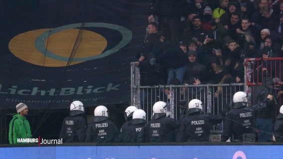 Ein Trupp an mit Helmen ausgerüsteten Polizei-Einsatzkräften steht einem randalierenden, schwarzgekleideten Fan-Block auf einer Tribüne gegenüber. © Screenshot 