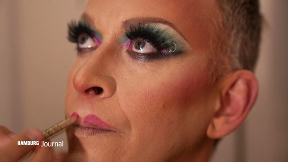 Ein Mann wird von einer Hand mit aufwendigem Make-Up geschminkt. © Screenshot 
