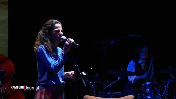 Auf einer spärlich beleuchteten Bühne singt eine Frau bei einem Auftritt in ein Standmikrofon. © Screenshot 
