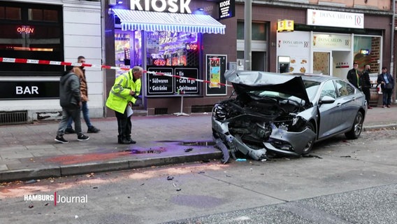 Nach einem Verkehrsunfall steht ein Auto mit komplett demolierter Front halb quer auf dem abgesperrten Bürgersteig einer belebten Straße. © Screenshot 