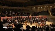Im großen Saal der Hamburger Elbphilharmonie treten bei einem Konzert viele hundert Menschen als großes Vokalensemble auf. © Screenshot 