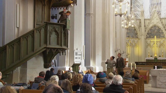 In einer Kirche spielen Darstellende ein Theaterstück in dem ein Schauspieler von der Kanzel spricht. © Screenshot 