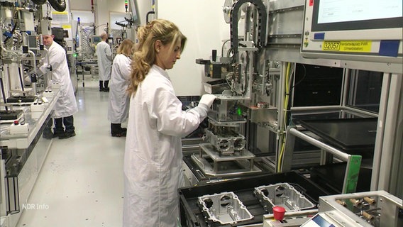 In einem Labor arbeiten mehrere Menschen in weißen Kitteln an Maschinen. © Screenshot 