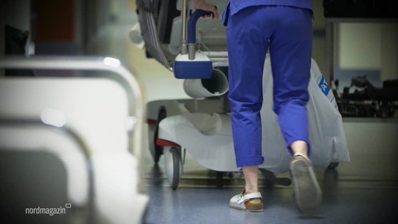 Eine Pflegekraft in blauer Arbeitskleidung schiebt ein Krankenhausbett über einen Flur. © Screenshot 