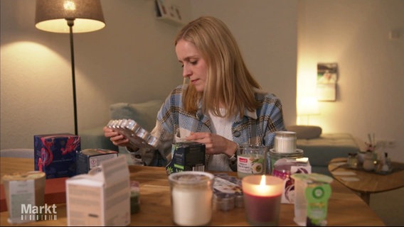 Eine junge Frau hält eine Packung mit Teelichten in der Hand; im Vordergrund unscharf zu sehen sind verschiedene Kerzen sowie Duftstäbchen. © Screenshot 