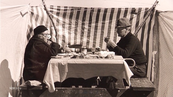 Eine Schwarzweißfotografie des Straßenfotografen Achim Sperber: Zwei Personen sitzen draußen an einem Tisch und essen, im Hintergrund weht eine gestreifte Plane. © Screenshot 