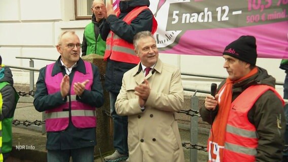 Mehrere streikende Personen in Warnwesten stehen vor einem Gebäude. © Screenshot 